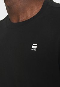 Tricou negru din bumbac, cu decolteu rotund și un logo alb "RAW" pe partea stângă a pieptului. Materialul pare neted, cu o ușoară strălucire.