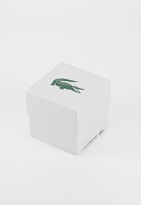 Baltas stačiakampis dėžutė su tekstūruota paviršiumi, ant viršaus vaizduojama tamsiai žalia Lacoste krokodilo logo, o šone - prekės ženklas.