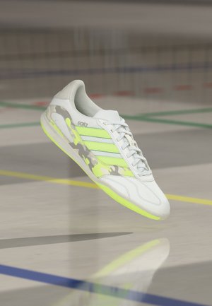 Weißer und neongrüner Sportschuh mit Camouflage-Details, der über einem Hallenboden mit farbigen Linien schwebt.