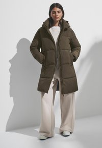Manteau matelassé vert olive avec capuche, doté d'une fermeture éclair sur toute la longueur et de deux poches avant. Porté sur un pantalon large beige clair.