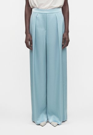 Femme portant un pantalon en satin bleu clair taille haute à jambes larges et des chaussures blanches pointues, debout devant un fond uni.