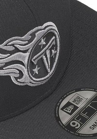 Casquette noire en tissu avec une finition texturée. Présente un logo en fil argenté avec des flammes et des étoiles. Inclut un autocollant New Era sur le bord.