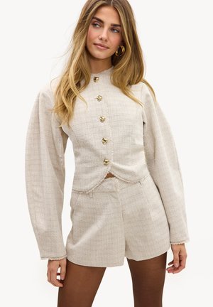 Jeune femme portant un haut boutonné à manches longues texturé couleur crème avec des manches bouffantes et un short taille haute assorti, boucles d'oreilles créoles dorées.