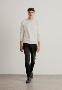 Suéter blanco de canalé, jeans negros ajustados y botas de cuero negras. El modelo se encuentra de pie contra un fondo liso, mostrando las proporciones del atuendo.
