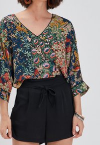 Top w kwiatowy wzór z dekoltem w serek, czarne szorty z wiązanym pasem. Top ma żywe kolory na ciemnym tle.