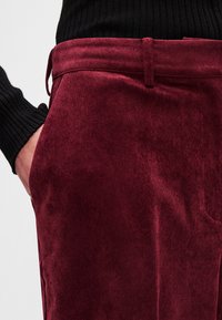 Burgundijas krāsas korduroja bikses ar gludu tekstūru, piedāvājot pieguļošu piegriezumu un sānu kabatu, kombinētas ar ribotu melnu džemperi.