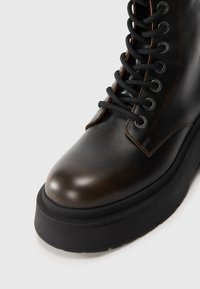 Bottine en cuir brun avec un bout arrondi, des lacets noirs, des œillets en métal et une semelle épaisse et texturée noire.