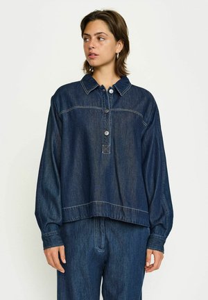 Denim-paitassa on kauluksellinen kaula, napitus etuosa ja roikkuneet olkapäät. Tumma sininen, jossa kontrastiväriset ompeleet. Kiristetty helma.