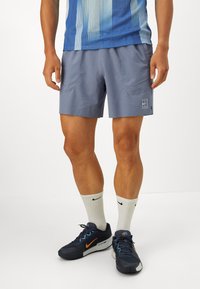 Shorts de sport gris avec poches latérales, coupe slim, et logo blanc. Associés à des baskets noires et orange et des chaussettes blanches arrivant au milieu du mollet.