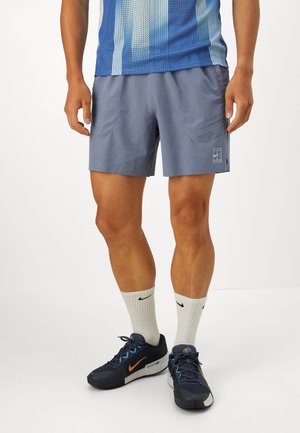 Shorts de sport gris avec poches latérales, coupe slim, et logo blanc. Associés à des baskets noires et orange et des chaussettes blanches arrivant au milieu du mollet.