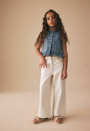 Jauna meitene ar gariem cirtainiem matiem, valkājot bezrokas džinsa topu, platām baltām biksēm un bēšām sandales, stāv vienkārša bēša fona priekšā.