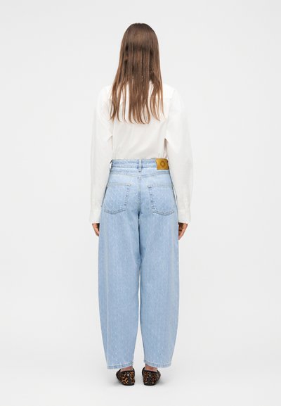 Άτομο με μακριά καστανά μαλλιά, φοράει λευκό πουκάμισο, ανοιχτό μπλε τζιν ψηλοκάβαλο με φαρδιά πατζάκια και μοκασίνια με λεοπάρ σχέδιο, γυρισμένο με την πλάτη.