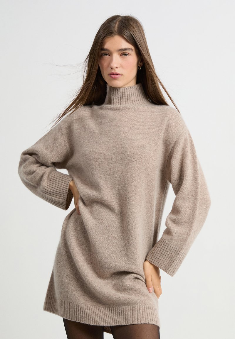 Vestido de punto beige con un alto cuello acanalado, mangas sueltas y puños acanalados, que presenta una textura suave y una silueta relajada.