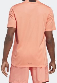 Camiseta atlética de color coral claro con cuello redondo y mangas cortas, confeccionada con tejido que absorbe la humedad y con un logotipo en el dobladillo inferior.