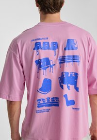 Rosa t-shirt med kort ärm som har blå grafiska tryck av olika stoldesigns och texten "THE SUNDAY CLUB" på baksidan.