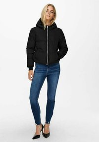 Doudoune noire avec capuche, dotée d'une fermeture éclair sur le devant et de poches latérales, portée par-dessus un jean skinny bleu et des talons aiguilles noirs.