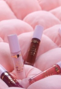 Vier Lippenprodukte mit sanfter Pastellverpackung, die auf einem samtigen rosa Hintergrund präsentiert werden. Auffällige Farben sind Hellrosa, Braun, Dunkellila und Koralle.