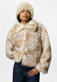 Giacca cropped in pelliccia sintetica beige con macchie scure, dotata di un ampio colletto e texture morbida. Abbinata a un cappello fluffy coordinato.