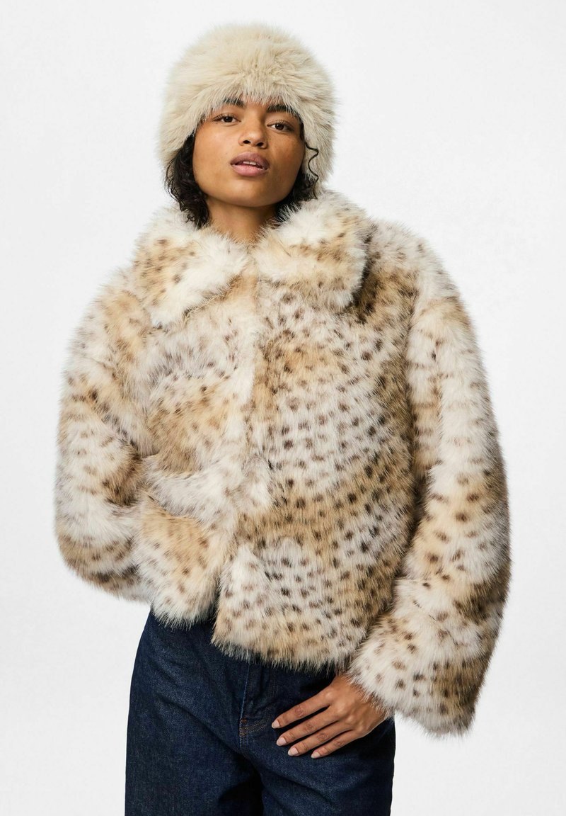 Giacca cropped in pelliccia sintetica beige con macchie scure, dotata di un ampio colletto e texture morbida. Abbinata a un cappello fluffy coordinato.