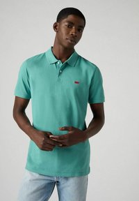 Turkos pikétröja i bomull med två knappslå, korta ärmar och en liten röd logopatch på vänster bröst. Ljus denimjeans.