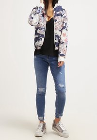Donna che indossa una giacca bomber con motivo floreale, top nero, jeans attillati strappati e sneakers metalliche, in piedi contro uno sfondo semplice.