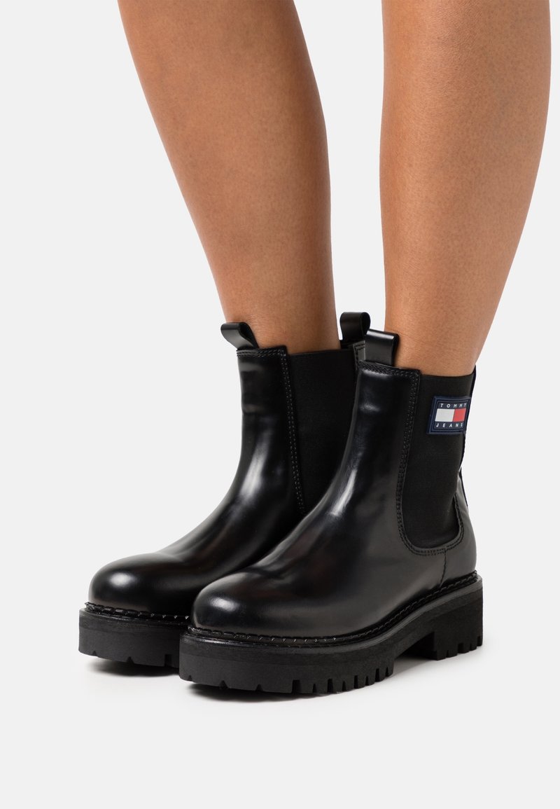 Bottines en cuir noir avec semelles épaisses à crampons portées sur jambes nues, dotées de panneaux élastiques sur les côtés et d'un patch de marque sur le côté extérieur.