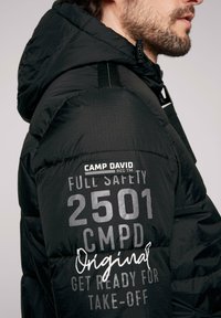 Chaqueta negra acolchada con capucha, que presenta varios estampados y logotipos reflectantes en las mangas, tela texturizada y un diseño ajustado.