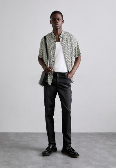 Chemise à carreaux noir et blanc à manches courtes, débardeur blanc, pantalon noir, chaussures noires, tenant un sac noir, debout contre un fond gris.