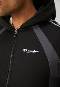 Nærbilde av en person som har på seg en svart hettejakke med glidelås og grå skulderdetaljer, og et Champion Products Inc.-logo på brystet.