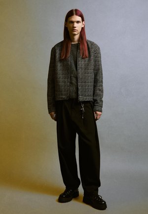 Schwarz-weiße gemusterte Bomberjacke, grauer Pullover darunter, lockere schwarze Hose und klobige schwarze Schuhe. Fließende Texturen und entspannte Passform.