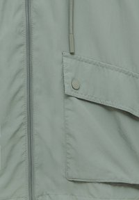 Lysegrøn jakke i glat stof med fuld lynlås, stor klaplomme med trykknapdetalje og snøre ved halsen.
