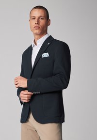 Blazer navy con dos bolsillos delanteros, un cuello con muescas y un acento de cuatro botones en los puños. Una camisa blanca y un pañuelo de bolsillo con patrón complementan el look.