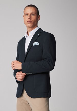 Blazer marine avec deux poches avant, un col cranté et un accent de quatre boutons sur les manches. Chemise blanche et pochette à motifs complètent le look.