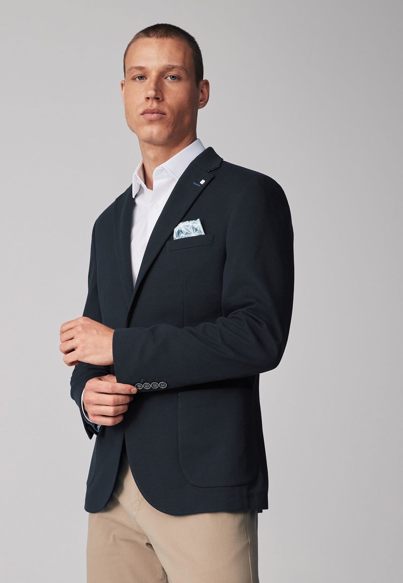 Blazer navy con dos bolsillos delanteros, un cuello con muescas y un acento de cuatro botones en los puños. Una camisa blanca y un pañuelo de bolsillo con patrón complementan el look.