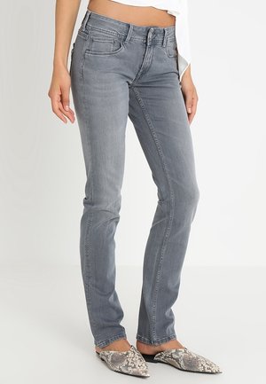 Kobieta w szarych, dopasowanych jeansach o średnim stanie oraz w butach wsuwanych w wężowy wzór, stojąca na jasnym, jednolitym tle.