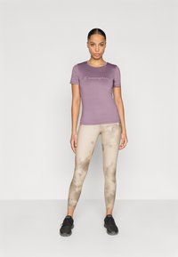 Μωβ κοντομάνικη μπλούζα με το λογότυπο "Champion", συνδυασμένη με μπεζ κολάν tie-dye και μαύρα αθλητικά παπούτσια. Ομαλό ύφασμα και εφαρμοστή σχεδίαση.