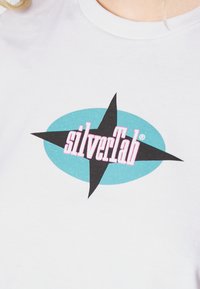 Bílý bavlněný tričko s tyrkysovým oválem, černou hvězdou a slovem "silverTab" v růžovém písmu.
