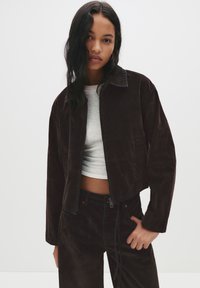 Brahkaste hlače iz kordura s svetlo modrim šivom, v kombinaciji s sivo majico. Model pozira z rokami v žepih, kar prikazuje ujemanje oblačila.