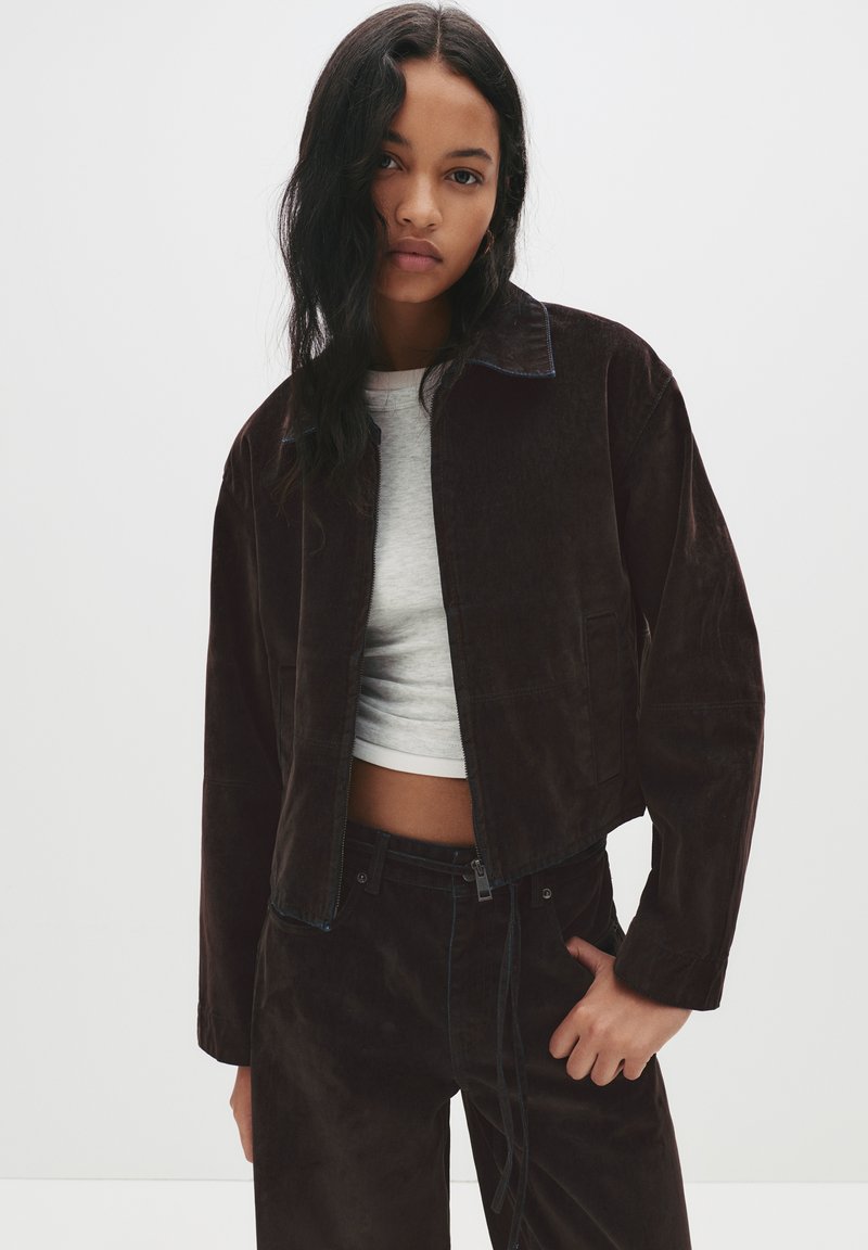 Brahkaste hlače iz kordura s svetlo modrim šivom, v kombinaciji s sivo majico. Model pozira z rokami v žepih, kar prikazuje ujemanje oblačila.