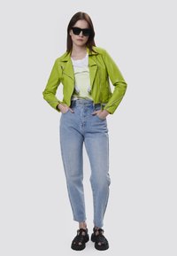 Giubbotto in pelle biker verde neon, t-shirt grafica bianca, jeans a vita alta blu chiaro e sandali chunky neri.
