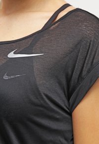 Musta urheilupaita, tehty kevyestä kankaasta, jossa on pyöreä pääntie, lyhyet hihat ja hopeanvärinen Nike-logo rinnassa.