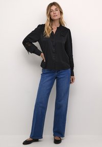 Blusa negra de manga larga con detalles de encaje en los puños y el collar, combinada con jeans de pierna ancha en azul claro y bailarinas negras. Textura suave, con botones en la parte delantera.