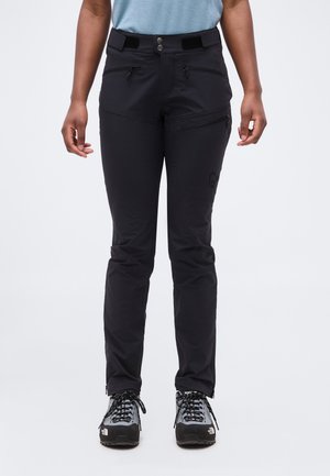 Άτομο που φοράει μαύρο παντελόνι εξωτερικού χώρου με πολλές τσέπες με φερμουάρ και μαύρα-γκρι παπούτσια πεζοπορίας, όρθιο σε λευκό φόντο.