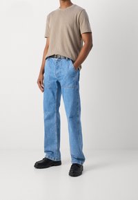 Uomo che indossa jeans straight leg blu chiaro, t-shirt beige a maniche corte, cintura nera e scarpe chunky nere su sfondo bianco.