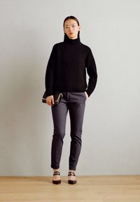 Pulover negru cu guler înalt și nervuri, pantaloni gri ajustați, pantofi negri cu accente decorative, model ținând o mică poșetă neagră.