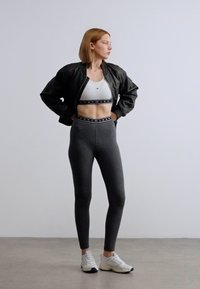 Μαύρο μπουφάν bomber πάνω από λευκό crop top, συνδυασμένο με γκρι κολάν ψηλόμεσα. Η εμφάνιση περιλαμβάνει ελαστικές λωρίδες με λογότυπο και λευκά αθλητικά παπούτσια.