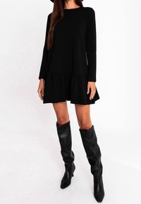 Czarna sukienka z długim rękawem i falbaną na dole, wykonana z miękkiego materiału. Zestawiona z czarnymi skórzanymi botkami do kolan oraz minimalistycznymi dodatkami.