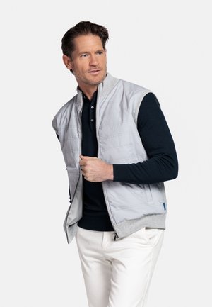 Man draagt een lichtgrijze doorgestikte bodywarmer over een zwart shirt met lange mouwen en witte broek, en sluit de rits van de bodywarmer terwijl hij opzij kijkt.