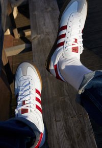 Vita idrottsskor med röda ränder, grå detaljer och en gummisul. Snörningen är vit och en logotyp är synlig på den yttre sidan.