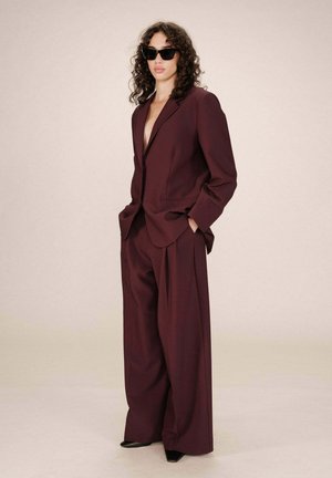 Femme avec des cheveux bouclés portant un costume oversize d'un prune foncé, des chaussures pointues noires et des lunettes de soleil noires, se tenant avec les mains dans les poches.